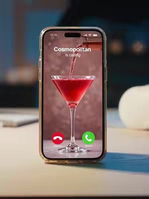 The image showcases an iPhone on a light wooden surface. The phone's screen displays a close-up of a cosmopolitan cocktail being poured into a stemmed glass. The background is blurred, suggesting a bar or dimly lit indoor setting. The phone screen features a calling interface with 'Cosmopolitan is calling' displayed, and the accept/decline call buttons. The phone is placed in a horizontal, eye-level shot, with a blurred background