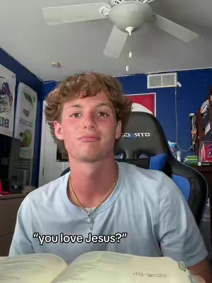 A young man with light brown curly hair is seated indoors, facing the camera directly. Text is layered over an open book. He wears a light blue t-shirt and a silver necklace. The background shows a room with a ceiling fan, posters, and a gaming chair. The lighting seems to be natural.