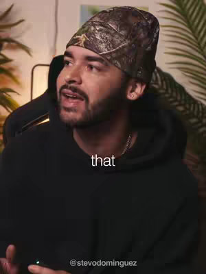 The image shows a man in a black hoodie and a camouflage beanie, looking towards the left of the frame and speaking. Text overlays the image.