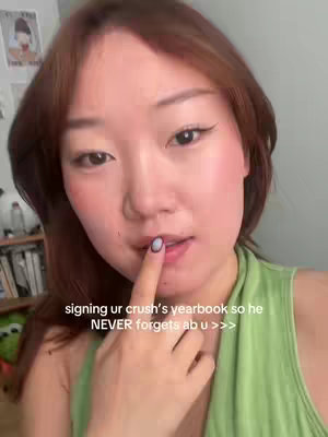 A selfie-style video featuring a young woman with long brown hair and light makeup. She is wearing a light green top and has one finger up to her lips, with a light blue nail. The background is a softly lit interior, with glimpses of bookshelves and other decor.