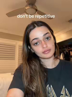 A woman with long brown hair and a black shirt looks directly at the camera with a neutral expression. The background includes a ceiling fan and the corner of a room.