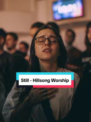 A woman with glasses and her eyes closed in worship in a room where other people in the background appear to be observing or participating in the worship, the text overlay indicates the song title and artist.