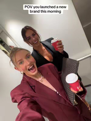 A selfie-style shot featuring two women, one in a burgundy blazer, the other in a black blazer, both holding red coffee cups. They are indoors, possibly in an office setting, with neutral-toned walls and a hint of a staircase visible in the background. The woman in the front wears a light-colored shirt beneath her blazer, and the woman in the back has a black fitted top. Both are smiling and appear excited.
