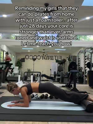 A woman is planking on a foam roller in a gym. She is wearing a black sports bra and leggings. The background has gym equipment and a large mirror. Text is overlaid, offering advice on how to do Pilates at home.