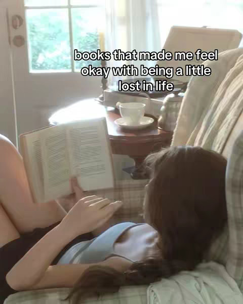 A person is lying on a couch with a book in hand. The person is dressed in a sleeveless top, with a view of a window, a side table with a teacup and a vintage record player with a lamp.