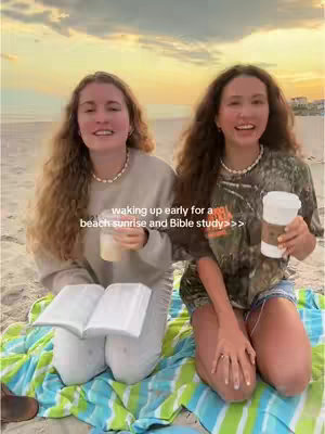 Two young women are on a beach at dawn, kneeling on a colorful blanket. The woman on the left is holding a clear cup with a beige liquid, while the woman on the right holds a coffee cup.  In the background you can see the ocean and sunrise colors.
