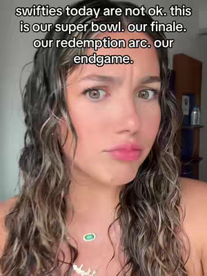 A selfie-style shot of a young woman with long, wavy hair and a neutral, concerned expression. She has fair skin, pink lips, and prominent eyebrows. Above her head, text in a clear font lists the following: swifties today are not ok. this is our super bowl. our finale. our redemption arc. our endgame. The background is blurred, but suggests a neutral indoor setting. There's a necklace in the shot.