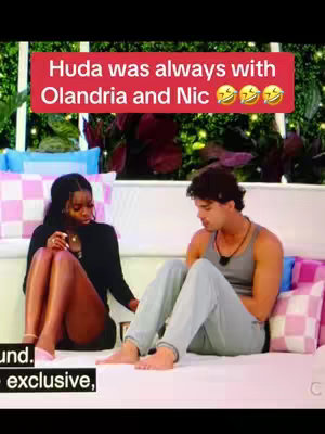 The image shows a scene from a television show with a red text overlay. Two people, a Black woman and a man, are seated on a white sofa adorned with various decorative pillows. The woman, dressed in a black outfit, is holding something in her hand and appears to be looking down; The man next to her, has his back to the sofa seat and is looking down as well.