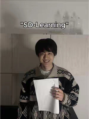 A young man with black hair smiles at the camera, holding a blank sheet of paper. He wears a patterned sweater over a white shirt, and the background shows a plain white wall with some bottles on a shelf.