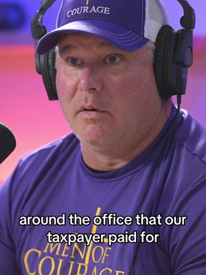 A man wearing a purple hat and shirt with a microphone in front of him. The background is blurred with pink and purple hues.