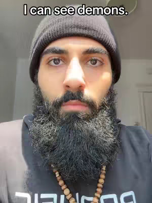 A man with a dark beanie, full beard, and a t-shirt with text is centered in the frame, staring directly at the camera. The background is softly lit with a hint of a window and wall.