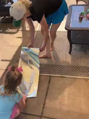A woman and a young girl are working together on a craft project outdoors. They are both standing on a tiled patio. The woman is bending over, holding a large, metallic sheet of material, and the girl is kneeling nearby, looking at the sheet. The composition captures a moment of collaboration and creativity.