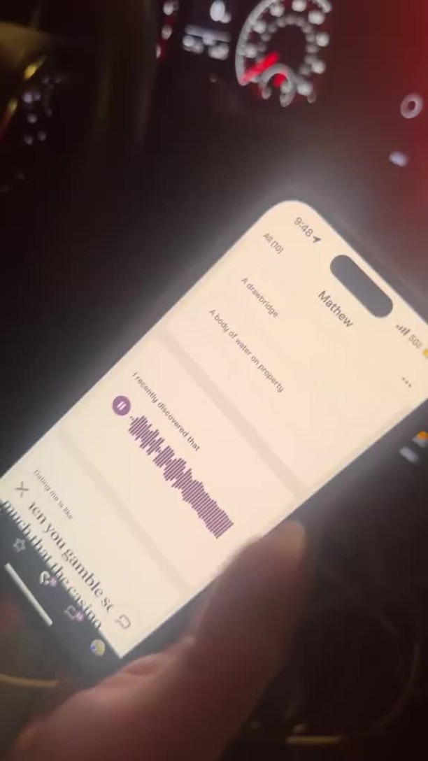 A person's hand holding a phone in a car interior. The phone screen is displaying a conversation thread with the name 'Mathew' at the top and an audio waveform related to a message. Other elements include a car dashboard and related driving instruments.