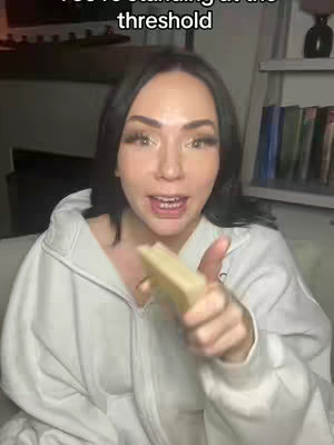 A woman with dark hair and makeup is holding a piece of bread toward the camera in a well-lit indoor setting. The text overlay, 'You're standing at the threshold' is at the top. Background elements include a shelf with books.
