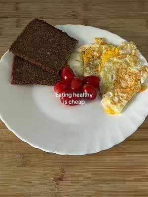 A plate with food on it on a wooden background. There are two slices of brown bread, a cluster of red cherry tomatoes, and two fried eggs with slight browning.