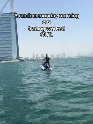 A man stands on a jet ski in the ocean. The Burj Al Arab hotel is visible in the background, along with a city skyline. The sky is blue and clear.