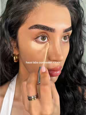 A close-up shot of a woman applying concealer near her eye. The skin is smooth and has a warm tone. A light-colored concealer is applied with a small applicator. The woman has dark, curly hair, and gold hoop earrings. The background is blurred, keeping the focus on her face.