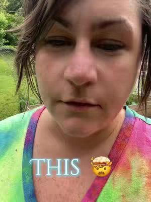 A close-up shot of a person's face. The person is looking down with a neutral expression. They are wearing a colorful tie-dye patterned shirt. The background is a blurred-out green scene, possibly a garden. Overlayed on the image is the word "THIS" in a light blue, glowing font, accompanied by a face-palm emoji.