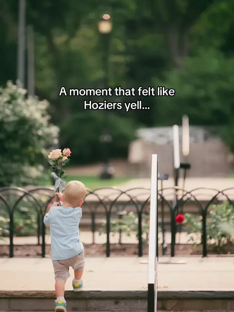 A young child, viewed from the back, walks up a set of stone steps, holding a bouquet of flowers. The steps are lined with a dark metal railing and a low wall. In the background, lush greenery, trees, and a lamppost provide a soft, blurred backdrop. Text overlayed on the image reads, 'A moment that felt like Hozier's yell...'