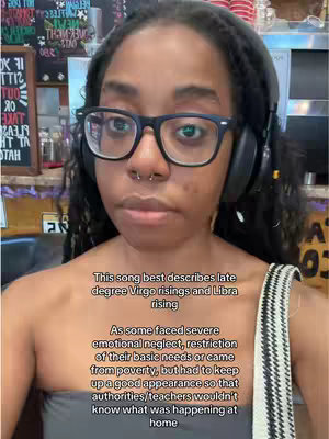 A person with dark hair and glasses looks into the camera, with text overlayed on the image. They are wearing headphones and a strap of a bag is visible on their shoulder. The background is blurry, likely in a cafe, with some text in the background appearing on a sign.