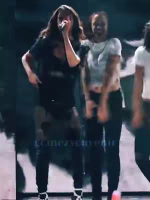 The image shows a live performance of a woman, presumably Selena Gomez, on stage. She is wearing a black t-shirt and black pants, and is holding a microphone. Two other people are in the background blurred. The scene is lit with stage lighting.