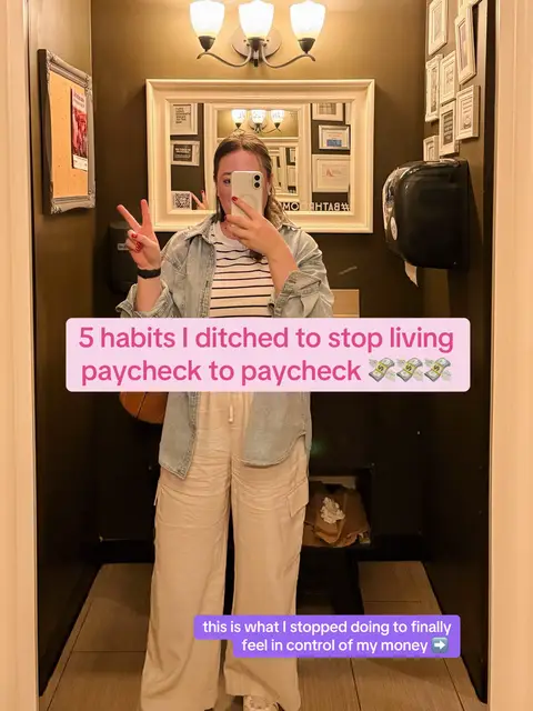 A selfie taken in a bathroom. A woman is in the center, holding a phone up to take the photo. She is wearing a striped shirt, a denim shirt, and tan pants. Text overlaid on the image says, "5 habits I ditched to stop living paycheck to paycheck". Additional text at the bottom says "this is what I stopped doing to finally feel in control of my money ->". The background includes a mirror, lights, and a bulletin board.