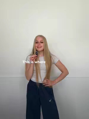 A young woman with long blonde hair is holding a microphone, smiling at the camera. She is wearing a white t-shirt and dark blue sweatpants. Text overlayed onto the image reads 'This is where'. The background is a plain white wall.