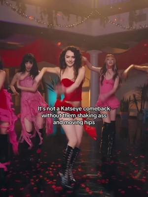 The image shows a group of women dancing in a nightclub setting. The central figure is wearing a red corset top and black shorts with high boots, dancing energetically. The background is a blurred mix of lights, red and pink decorations, and other dancers in similar outfits. The camera angle is low, capturing the dynamic movement of the dancers.