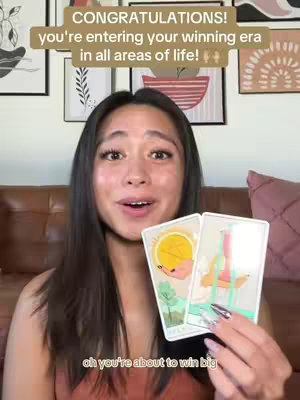 A woman is in the center of the frame, holding two tarot cards. She is smiling and looking directly at the camera, against a background of framed art and a brown backdrop. The video overlay features text.
