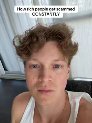 A close-up shot of a fair-skinned person with short, wavy, light brown hair, wearing a white tank top, looking directly at the camera. The background is blurred, but appears to be a room with curtains and a window.