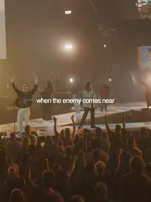 A concert stage is filled with performers and audience members with their arms raised. The stage is well-lit, and the background features a large screen and lighting rigs. Text overlayed on the image reads 'when the enemy comes near'.