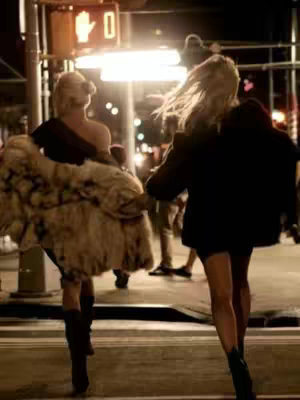 Two women walking on a city street at night. The woman on the left is holding a fur coat, while the woman on the right has her hair blowing in the wind. They are walking towards the center of the frame, away from the viewer with a street light in the background. The image has a dim, night-time aesthetic.