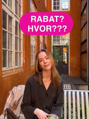 A woman with long blonde hair smiles in front of an orange building exterior. A pink oval with white text overlayed on top of her head reads: 'RABAT? HVOR???'. She is wearing a black top, and a beige coat and a purse are visible on the bench behind her.
