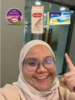 A woman with glasses and a headscarf smiles and points her finger towards the upper-right corner. Behind her, there are food ingredients like mascarpone, ladyfingers, and butter. In the background, there is a glass wall.
