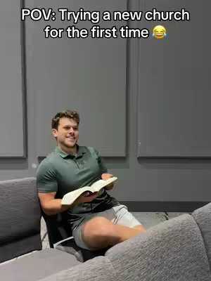 A man is sitting on a curved gray couch, holding a book open, smiling, in a room with gray sound-dampening panels on the wall. Text on the image says, "POV: Trying a new church for the first time 😅".