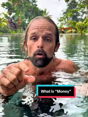 A man with a beard is submerged in water up to his chest, reaching a hand towards the viewer. A text overlay reads, 'What Is Money?' The background shows green foliage, suggesting a natural setting.