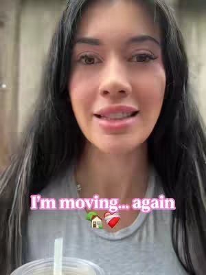 A selfie-style shot featuring a woman with long, dark hair and a light grey t-shirt. The woman is looking at something off-camera and smiling slightly. Overlayed on the screen is the text, "I'm moving... again," in a pink bubble letter style, interspersed with house and heart emojis.