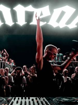 The cover image captures a nighttime music concert scene. In the foreground, a DJ setup is visible. A man with short hair wearing sunglasses is the central figure, captured with his arm raised. He is the focus of the composition and also has multiple tattoos on it. Beyond him, a crowd is visible, enhanced by the large, illuminated text in the background and dark atmosphere. The lighting adds to the energy of the concert.