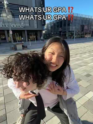 The photo shows two young women, one carrying the other piggyback style in front of a city setting. The woman on the bottom is holding the top woman's legs. The woman on top is smiling broadly, while the woman being carried is also smiling at the camera. The background shows a modern building made of glass and concrete, next to a building with a sign and an entrance with a VIP