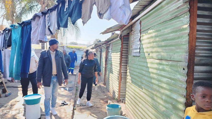 An outdoor shot captures a scene in an informal settlement, likely in Pretoria West, South Africa. Several corrugated iron shacks line a pathway. A man in a dark jacket, jeans, and a beanie leads, followed a woman in black athletic wear. Other individuals, including some in uniform, and a young child looking towards the camera, are also visible. Clothes hang on a line above, and buckets are scattered along the path. The lighting suggests a sunny day.