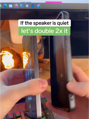 The cover image shows a person's hands holding two mobile phones. The phone on the left is angled towards the viewer, displaying its screen with an app icon. The phone on the right is facing away, with a black case. Above the phones, a text overlay in a white box reads "If the speaker is quiet let's double 2x it" in bold, green letters. The background is mostly blurred, but shows a wooden surface, some electronic devices, and a warm light source.