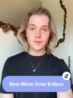 A medium shot of a young man with light hair and blue eyes. He's wearing a black shirt and a silver necklace. A text overlay at the bottom of the screen says 'New Moon Solar Eclipse.' The background includes a mirror and a wall, with a TikTok logo in the upper right corner.