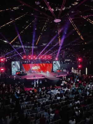 An overhead shot of a large, brightly lit event space, likely a church or conference hall. The stage is at the far end, with a large screen displaying a logo or image. The audience fills the seats, and there are spotlights and stage lights creating a dynamic atmosphere.