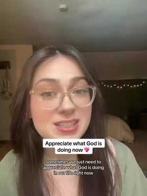 A woman with glasses and brown hair looks directly into the camera. Overlayed text in a white box reads "Appreciate what God is doing now", with another box below containing further text. The background is blurred, but includes part of a wall and a light.