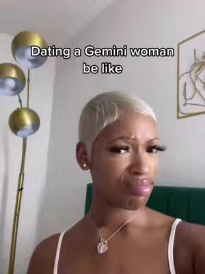 The image is a selfie of a woman with short, bleached blonde hair, pursing her lips. She has prominent eyelashes, simple earring, and a gold necklace. The background includes a floor lamp with gold globe shades and a glimpse of a wall and a framed artwork. Overlayed text states, 'Dating a Gemini woman be like'.