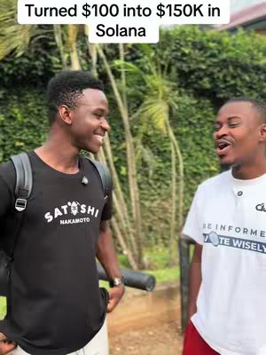 Two young men are standing side-by-side outdoors, with lush green foliage visible in the background. The man on the left is wearing a black shirt with white text and a backpack, while the man on the right is wearing a white shirt with text. The camera is angled slightly upward.