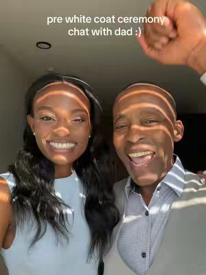 A selfie photo featuring a young Black woman and an older Black man smiling at the camera. The woman has long, dark hair and is wearing a light-colored top. The man is wearing a button-down shirt and a light vest. Both are smiling and the sun gives the photo a well-lit feel.