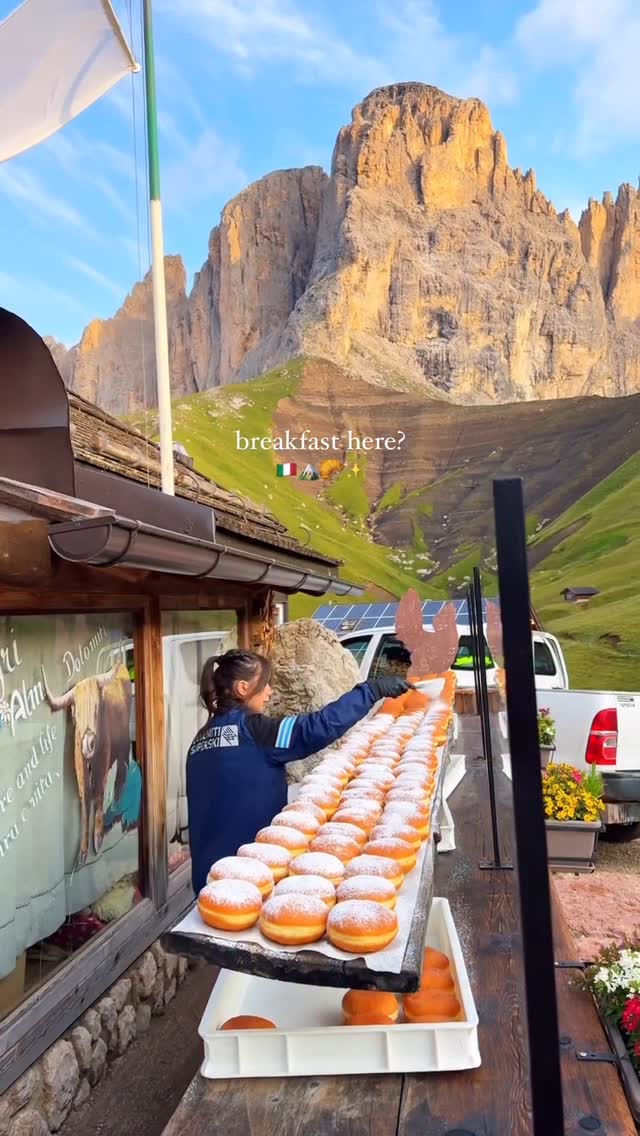 A woman in a blue jacket is arranging many freshly baked, powdered sugar-covered donuts on a wooden surface, with a mountain range in the background. The text overlay says "breakfast here?" with a series of icons (Italian flag, mountains, and sparkles).