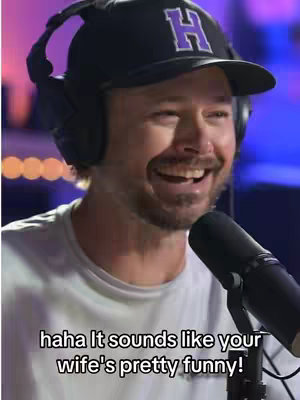 A man wearing a black cap with a white 'H' logo sits in front of a microphone, smiling and laughing. The image has a soft, blurred background.