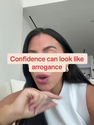 A woman with long, dark hair and a sleeveless white top is partially visible in the frame. A red rectangle covers her forehead, with the text 'Confidence can look like arrogance' displayed on it. She is holding her hand near her mouth, gesturing slightly. The background is blurred.